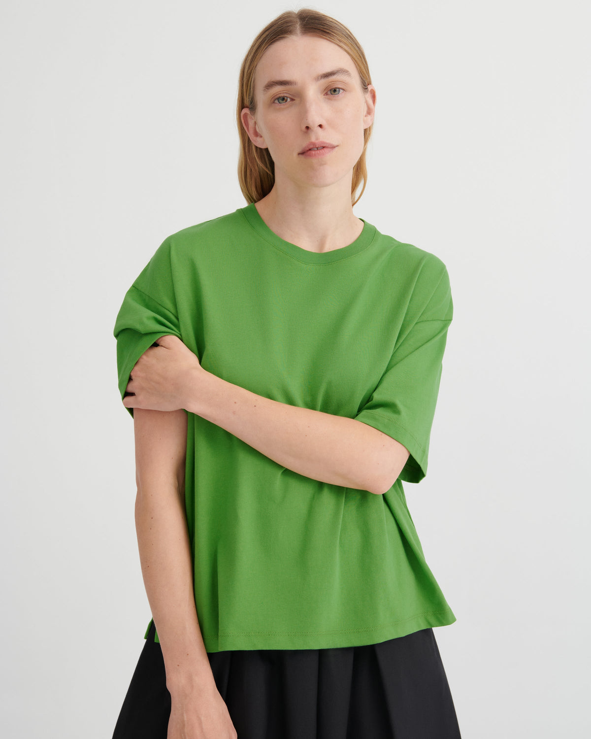 A woman wearing a bright green short-sleeve crewneck t-shirt and a black skirt, standing against a light gray studio background.