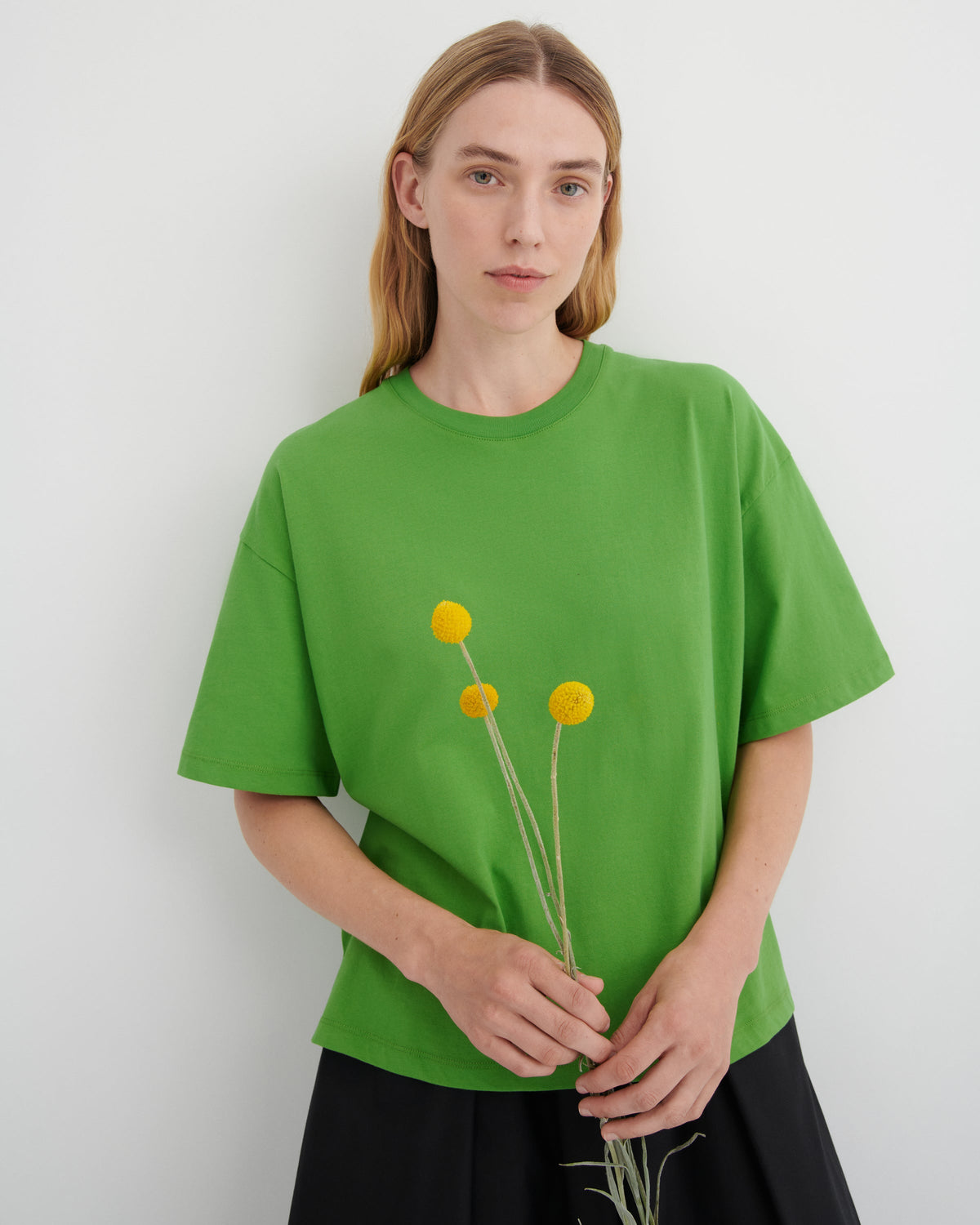 A woman wearing an oversized bright green t-shirt with a small yellow flower graphic on the front, paired with a black skirt, holding three yellow flowers against a light gray background.
