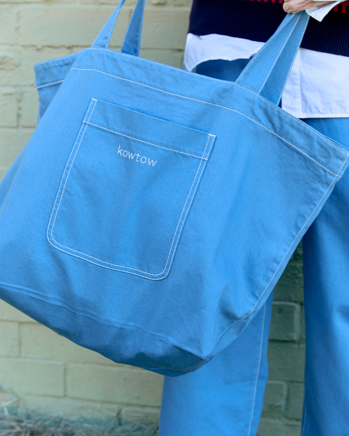A light blue canvas tote bag with white contrast stitching and a front pocket embroidered with the brand name 'kowtow', carried by a person wearing matching blue overalls against a light brick wall.