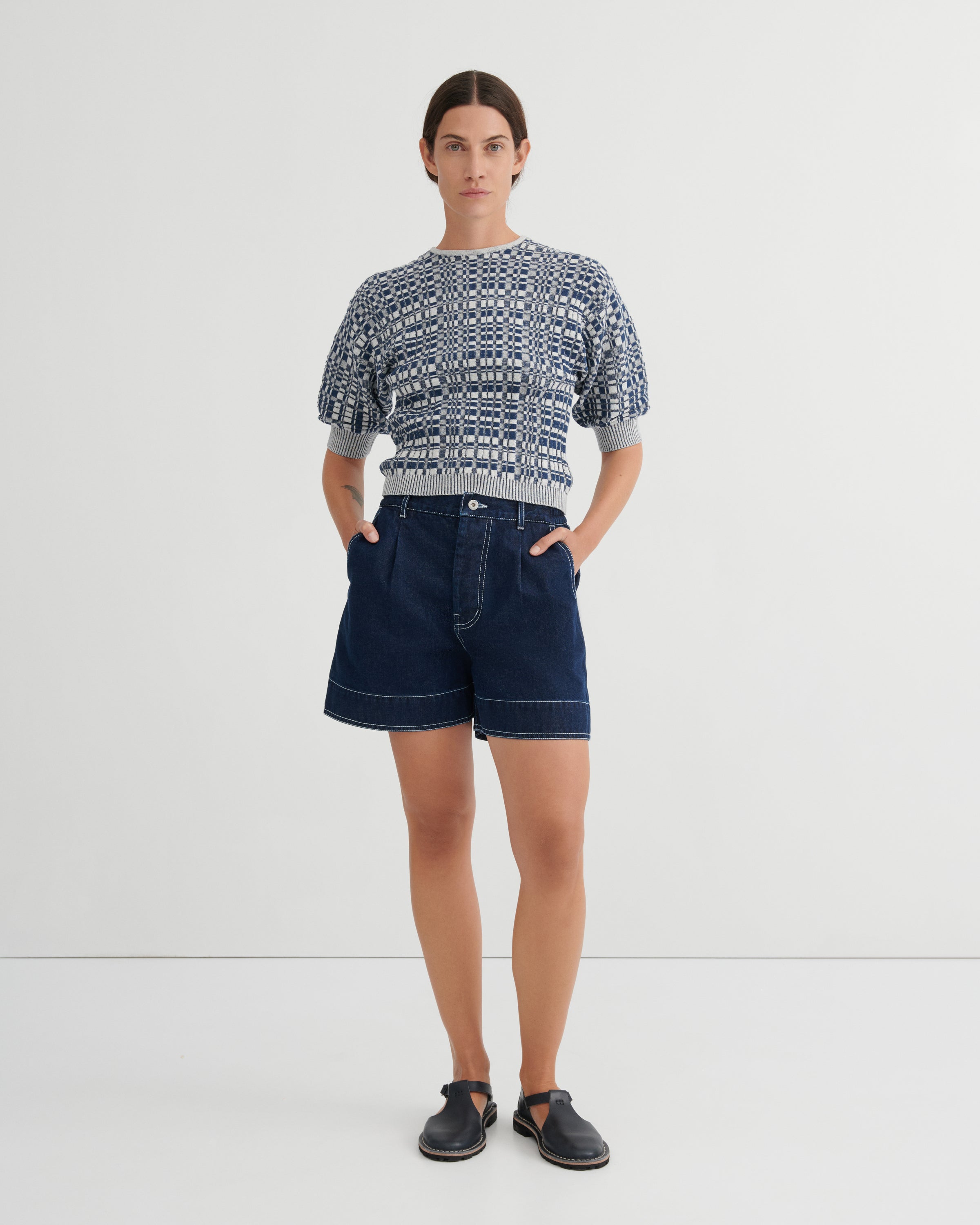 A model stands against a light gray studio background wearing a short-sleeve knit top with a blue and white geometric pattern, dark blue denim shorts, and dark slip-on shoes.