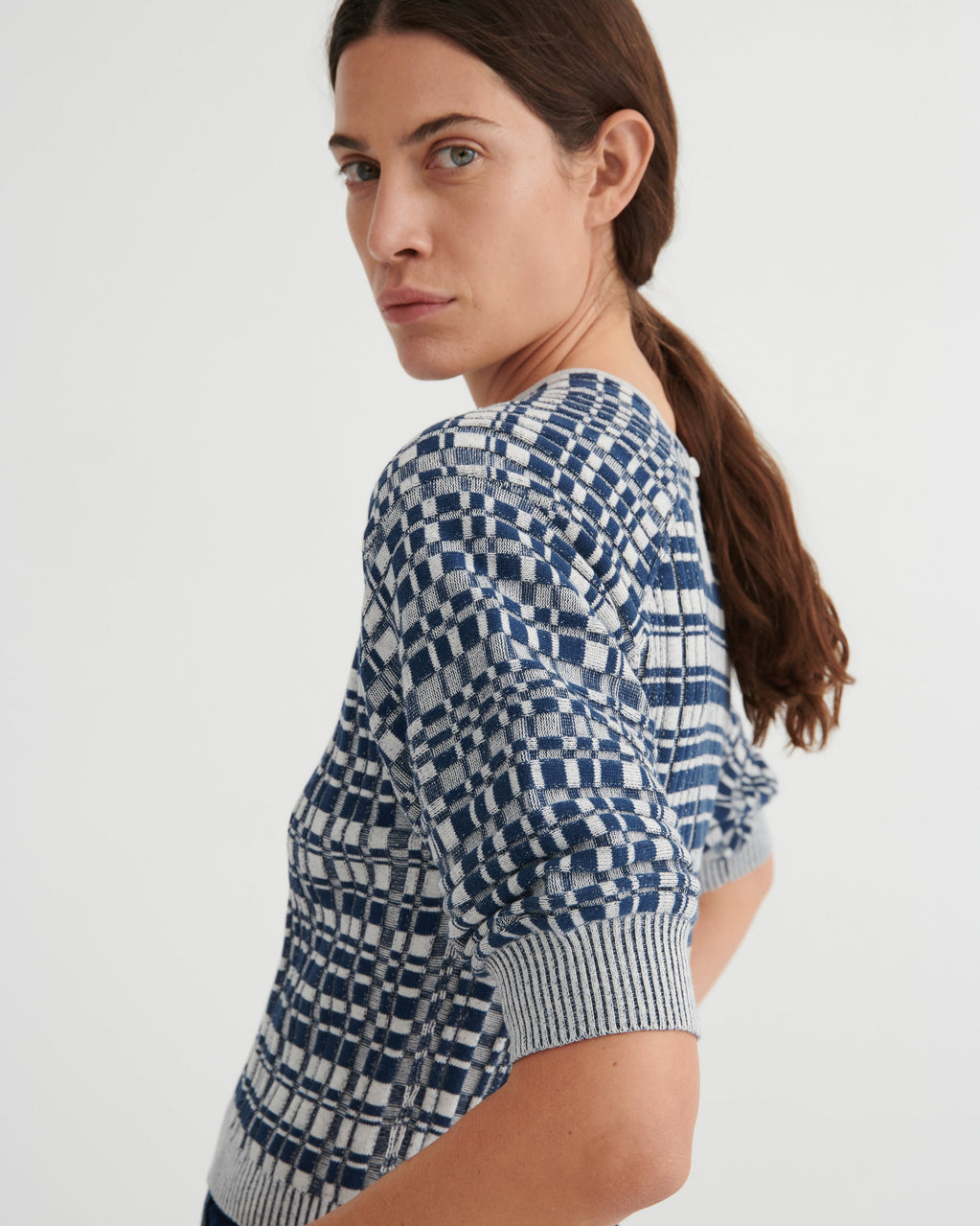 A woman wearing a short-sleeve knit top with a blue and white geometric checkerboard pattern, shown from a side-back angle against a plain light gray studio background.