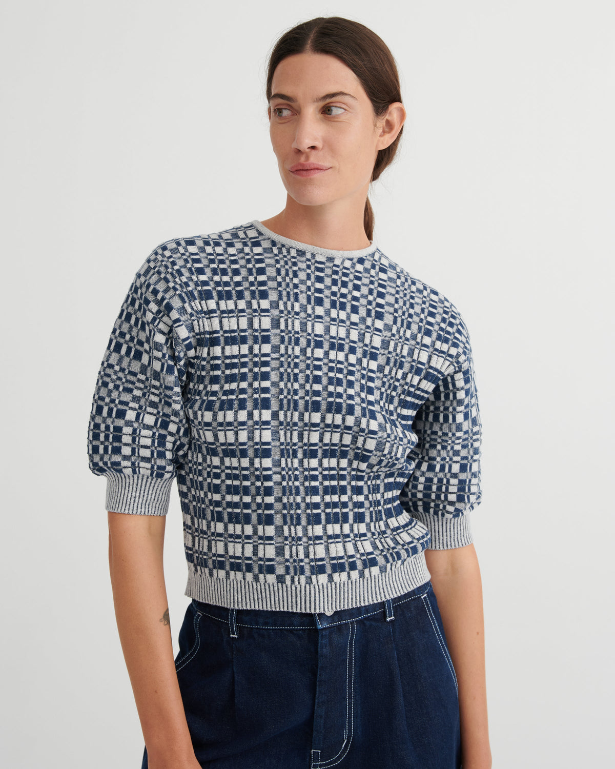 A woman wearing a short-sleeve knit top with a navy and white geometric grid pattern, paired with dark blue jeans, against a plain light gray studio background.
