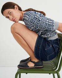 A woman crouches on an olive green metal chair wearing a blue and white patterned knit top, dark denim shorts, and black Mary Jane-style flat shoes against a plain light background.