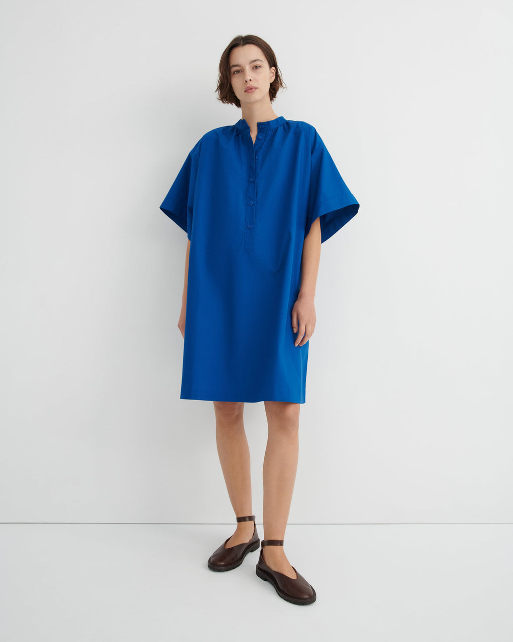 A woman modeling a loose-fitting, short-sleeved blue dress with a buttoned mock neck.