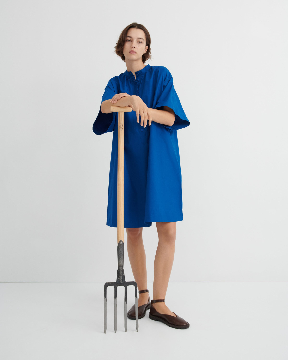 A model wearing a loose, short blue dress and dark brown shoes, holding a large pitchfork with a wooden handle.