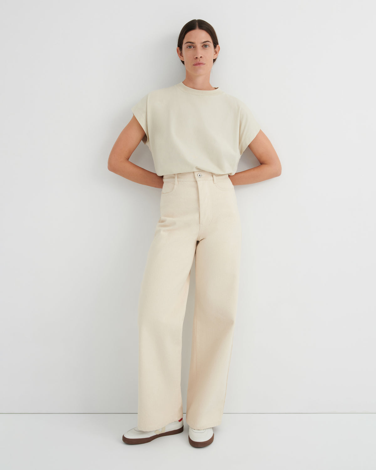 A woman modeling a light cream short-sleeve crewneck top and matching high-waisted wide-leg trousers, standing against a plain white studio wall.