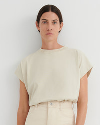 A woman wearing a cream-colored short-sleeve crewneck t-shirt and matching cream trousers, standing against a plain light gray wall.