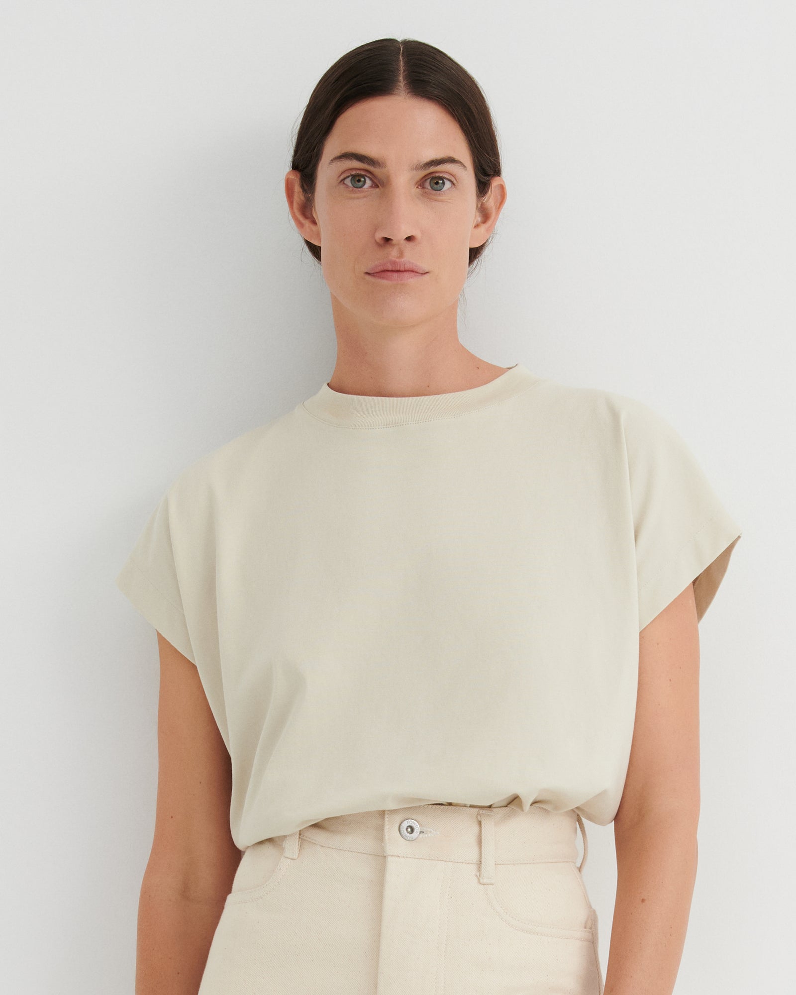 A woman wearing a cream-colored short-sleeve crewneck t-shirt and matching cream trousers, standing against a plain light gray wall.
