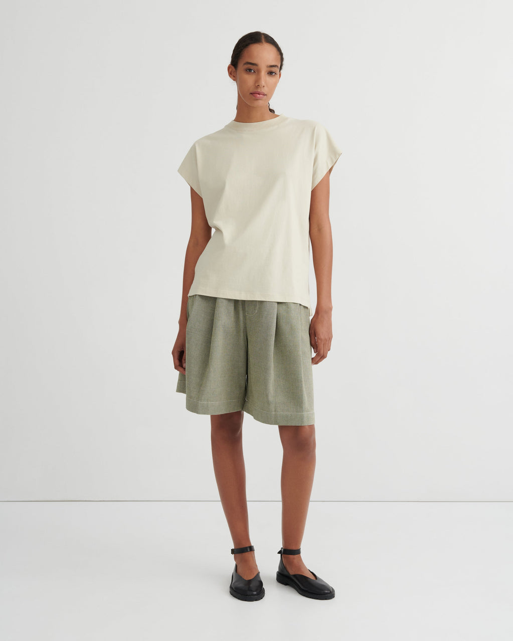 A model stands against a plain light gray studio background wearing a cream-colored short-sleeve t-shirt and olive green linen shorts, paired with black flat shoes.