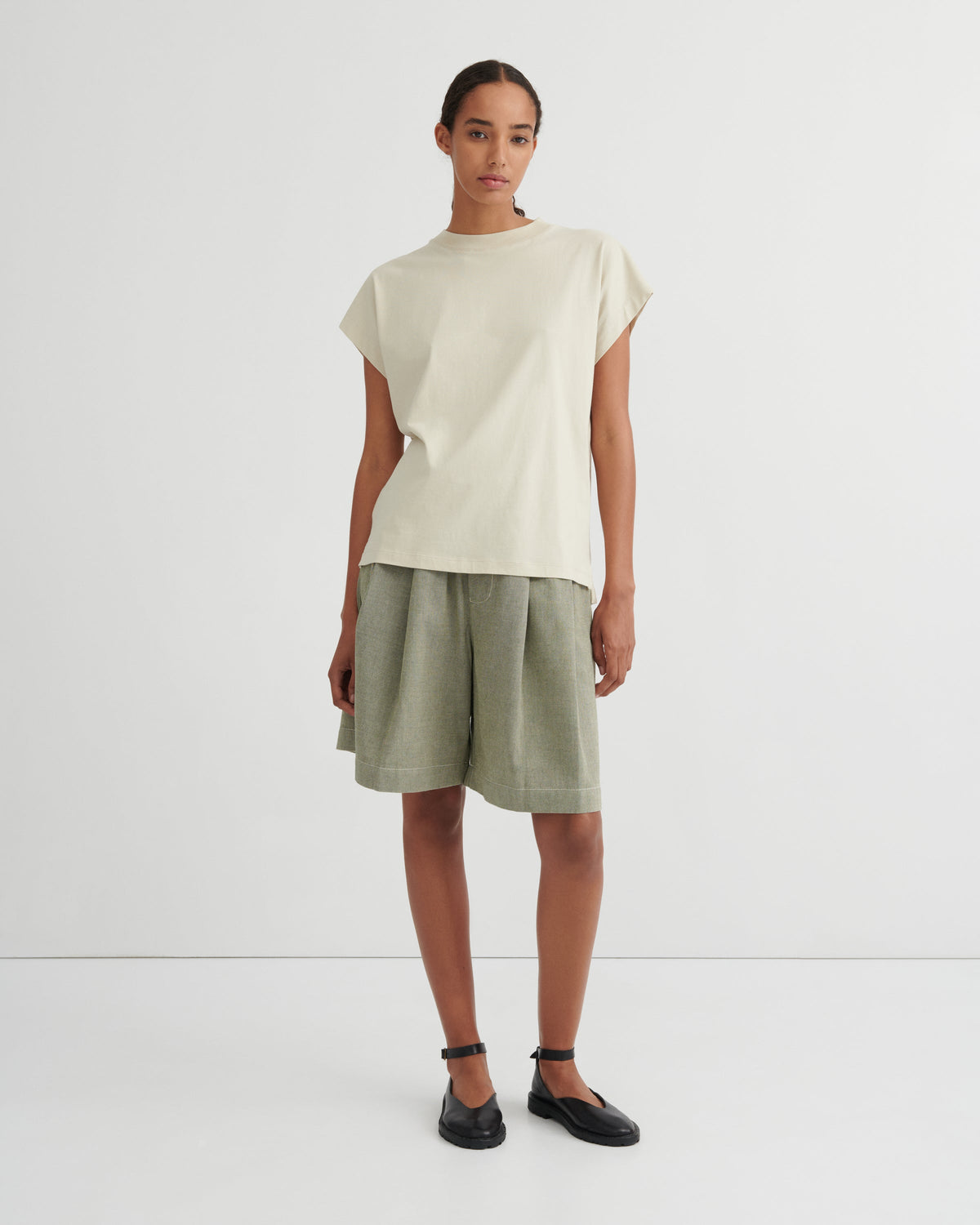 A model stands against a plain light gray studio background wearing a cream-colored short-sleeve t-shirt and olive green linen shorts, paired with black flat shoes.