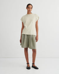 A model stands against a plain light gray studio background wearing a cream-colored short-sleeve t-shirt and olive green linen shorts, paired with black flat shoes.