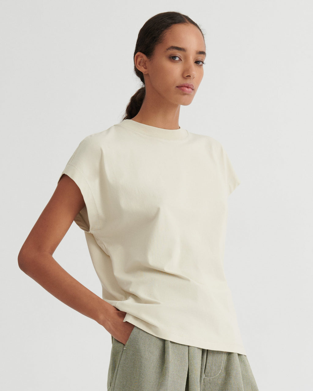 A woman modeling a cream-colored short-sleeve crewneck t-shirt, paired with light sage green trousers, against a solid light gray background.