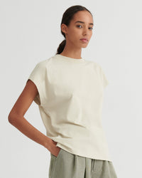A woman modeling a cream-colored short-sleeve crewneck t-shirt, paired with light sage green trousers, against a solid light gray background.