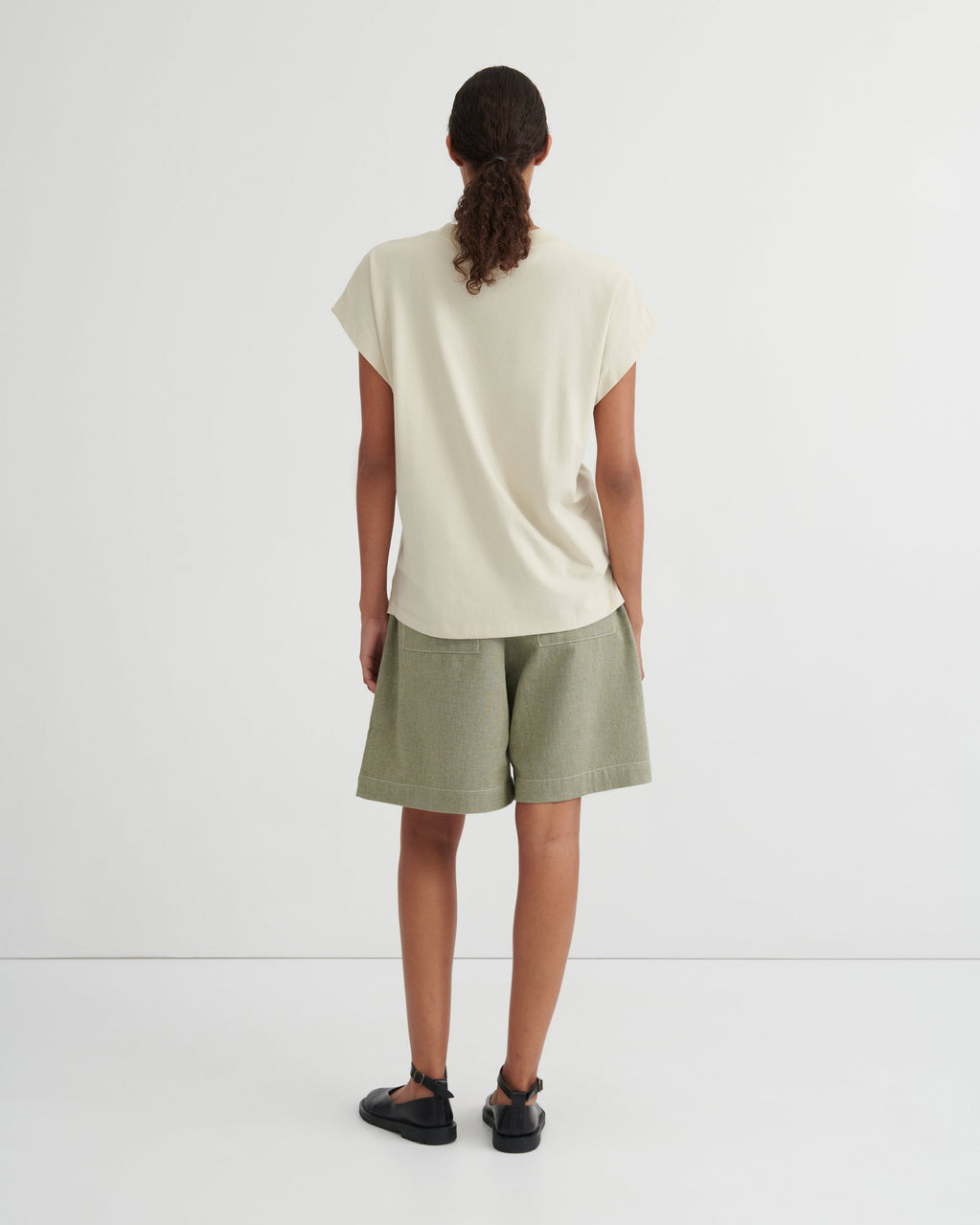 A model viewed from behind wearing a cream-colored short-sleeve t-shirt and sage green shorts against a plain white studio background.