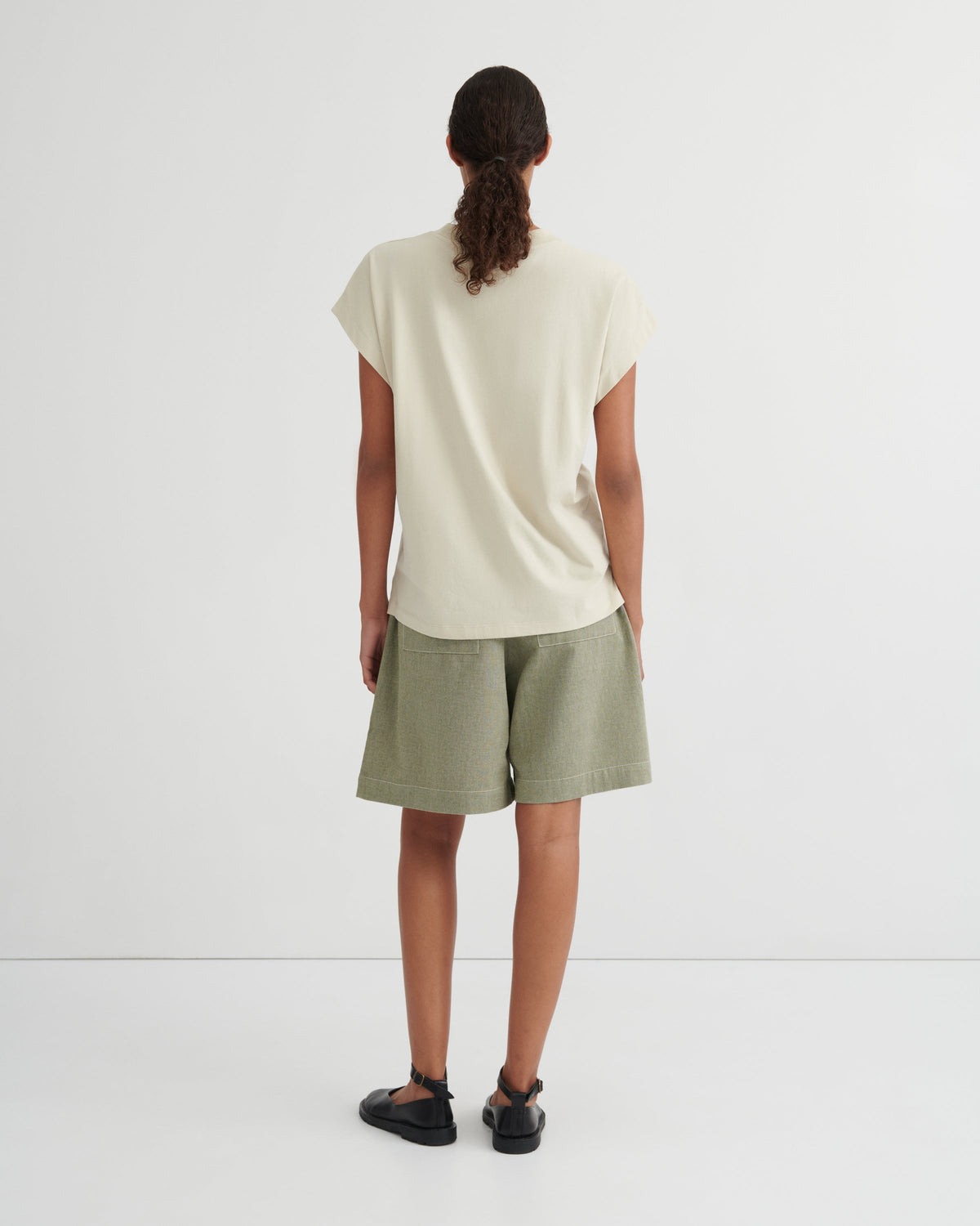 A model viewed from behind wearing a cream-colored short-sleeve t-shirt and sage green shorts against a plain white studio background.