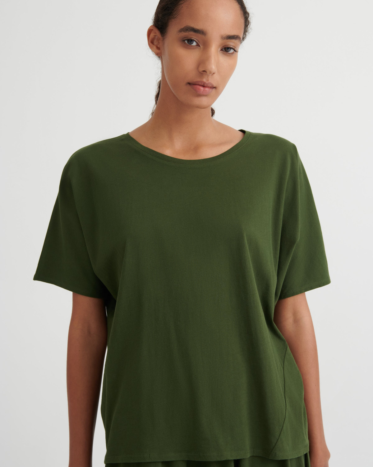 A woman wearing a loose, short-sleeve crewneck t-shirt in olive green, photographed against a plain light gray studio background.