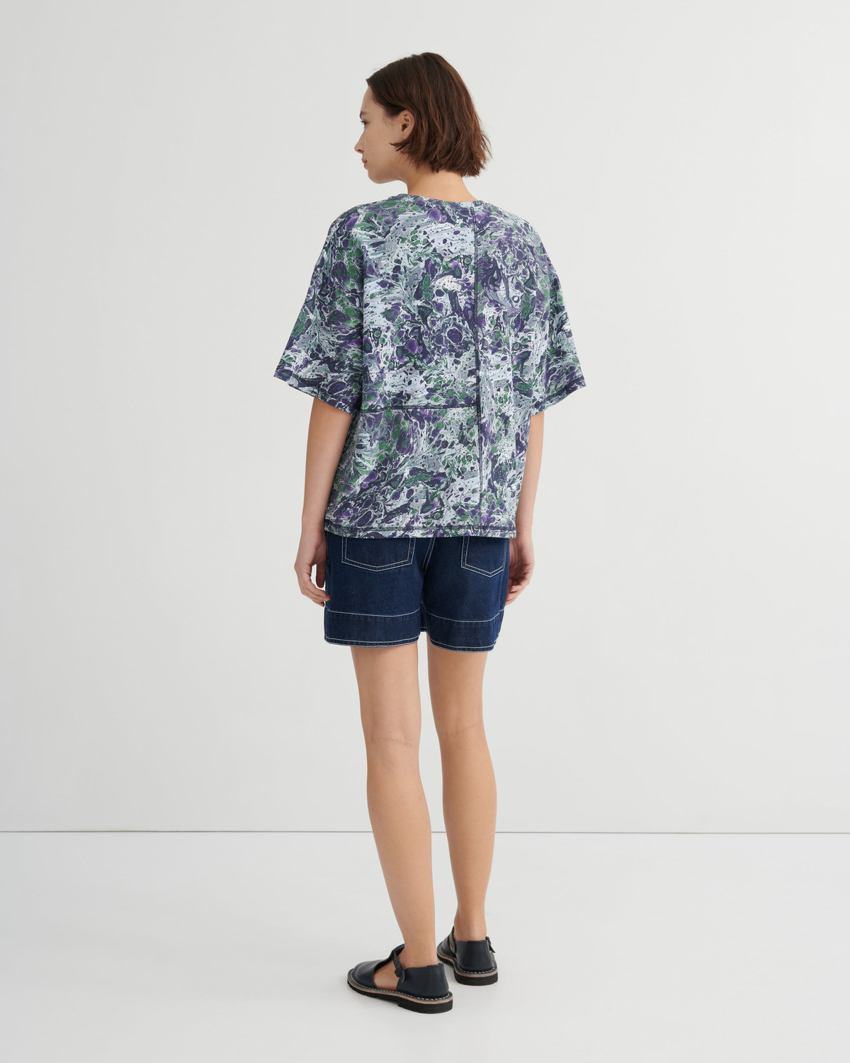 A woman is shown from the back wearing a short-sleeve patterned top with a blue and green marble print, paired with dark denim shorts and black sandals, against a plain white studio background.