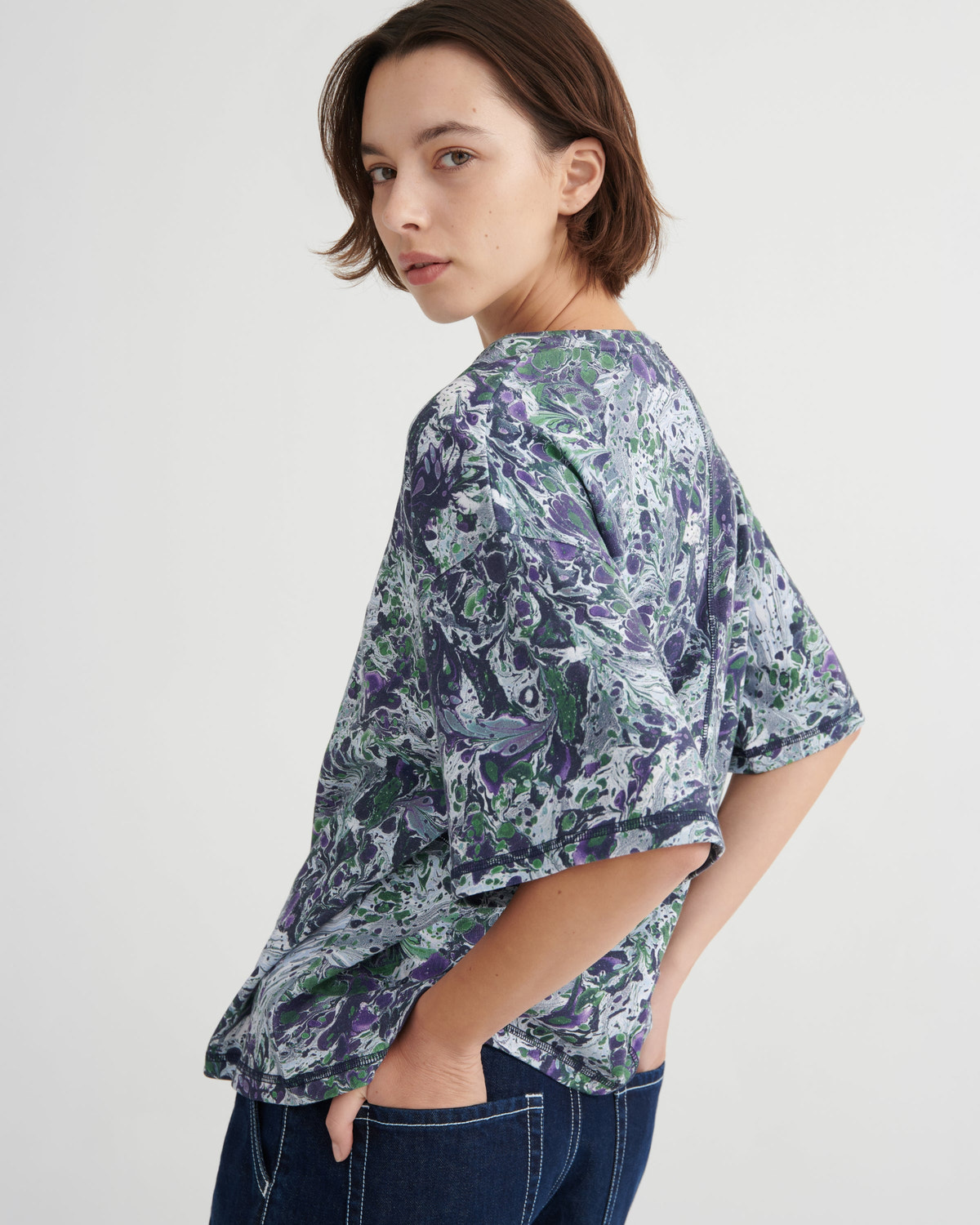 A woman wearing a loose short-sleeve top with a blue, purple, and green marble floral print, paired with dark blue jeans, photographed against a light gray studio background.