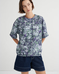 A woman modeling a short-sleeve button-front top with an abstract marble print in purple, green, and white, paired with dark denim shorts against a plain light gray studio background.