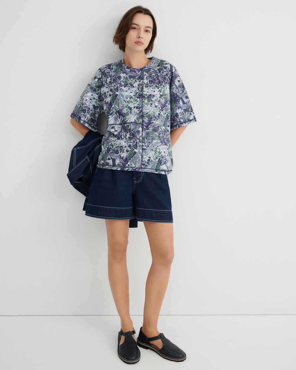 A model stands against a plain light gray studio wall wearing a short-sleeve button-front shirt with a blue, green, and white marble print, dark denim shorts, and black slip-on shoes. A dark blue jacket is draped over one shoulder.