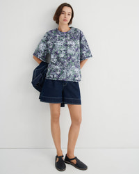 A model stands against a plain light gray studio wall wearing a short-sleeve button-front shirt with a blue, green, and white marble print, dark denim shorts, and black slip-on shoes. A dark blue jacket is draped over one shoulder.
