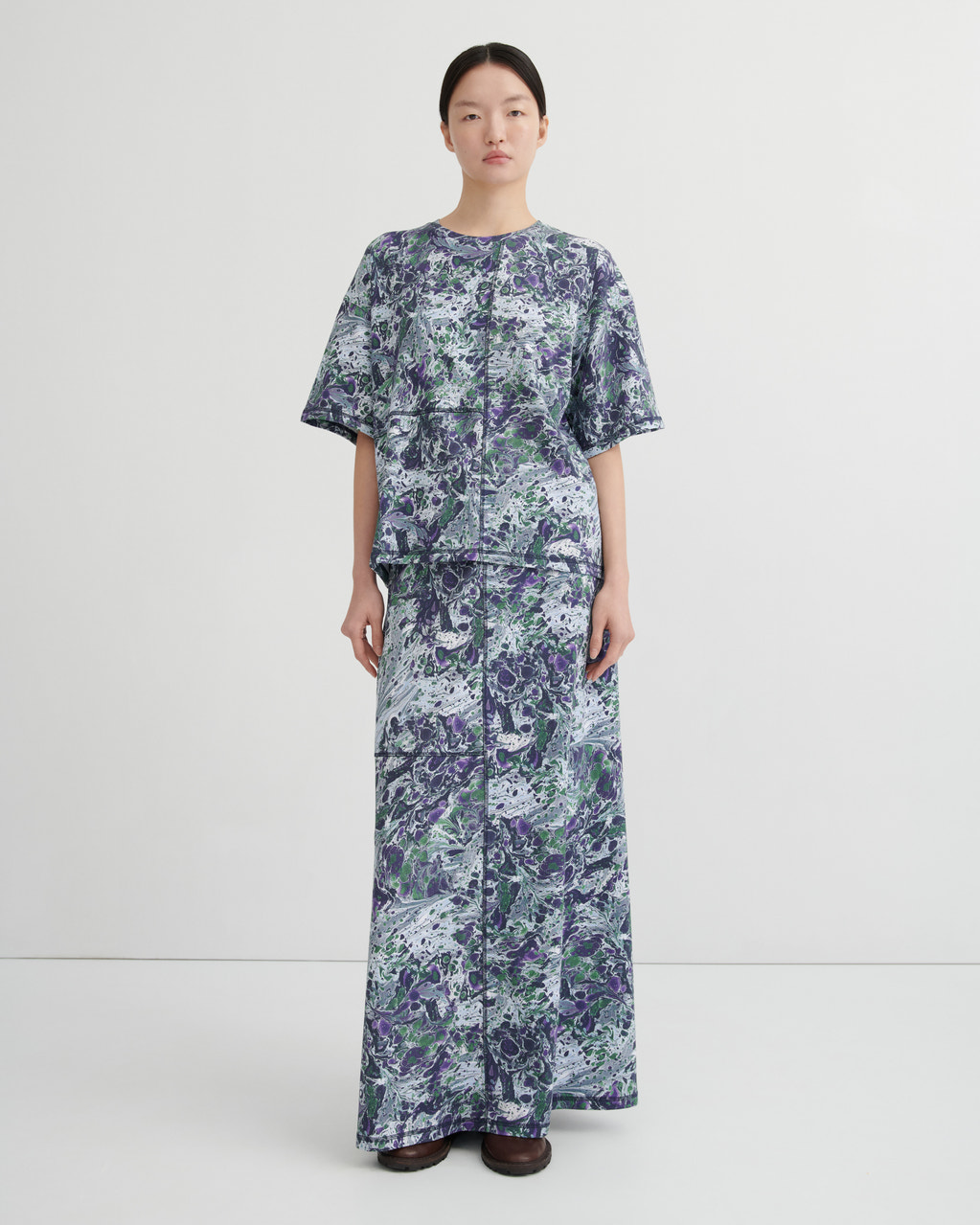 A model stands against a plain light gray studio background wearing a long-sleeved maxi dress with a blue, purple, and green abstract marbled pattern.