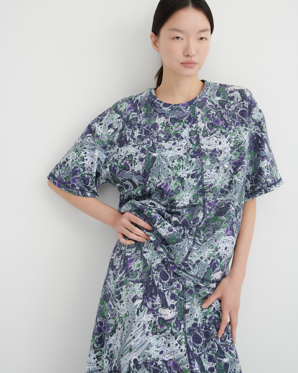 A model wearing a short-sleeve dress with a purple, green, and white abstract marble pattern, standing against a plain light gray wall.