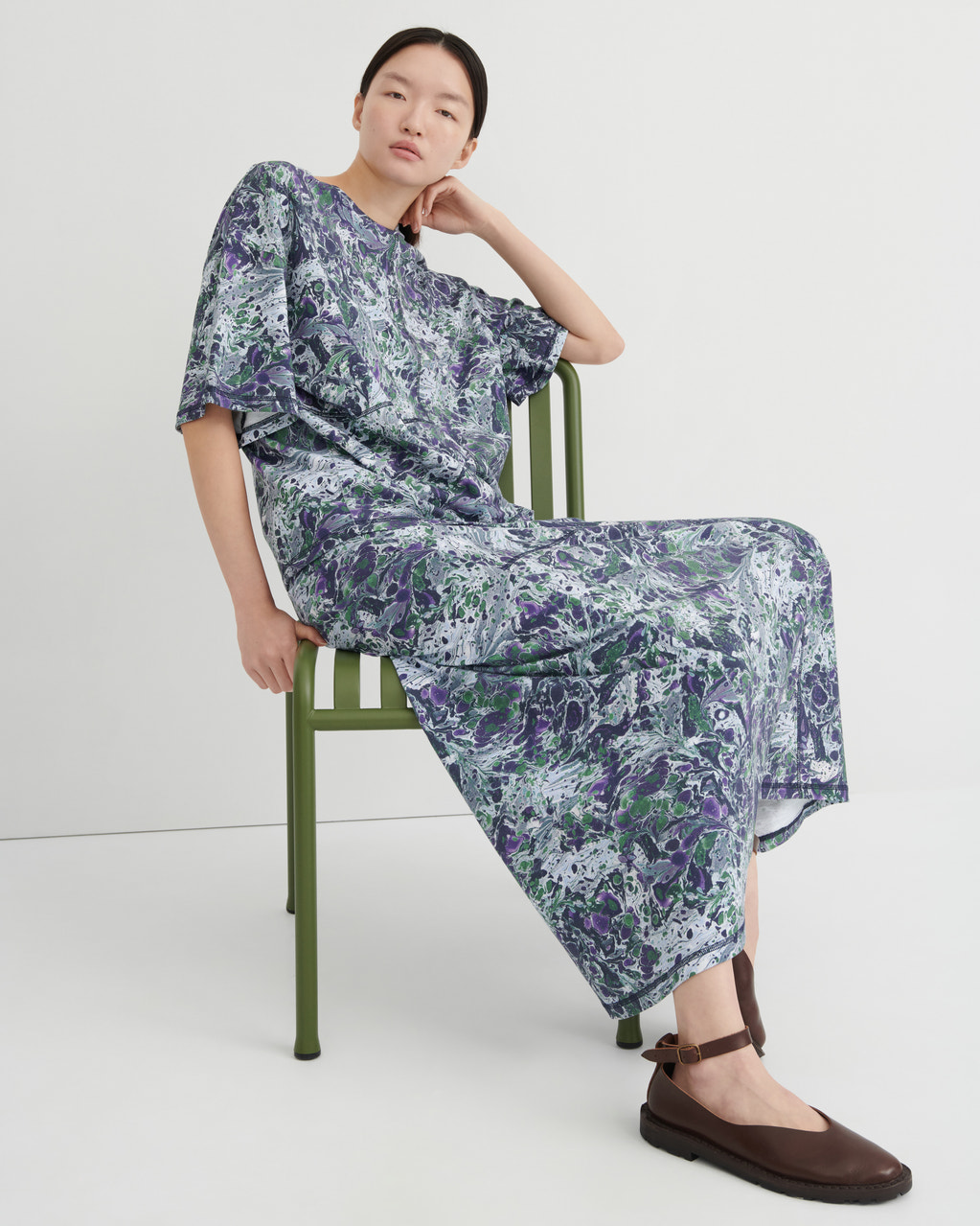 A model sits on a green metal chair wearing a loose, short-sleeved maxi dress with a blue, purple, and green abstract marble print, paired with dark brown flat shoes, against a plain white studio background.