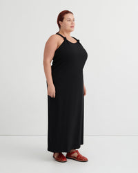 A woman modeling a black halter-neck maxi dress with shoulder cutouts, paired with brown leather sandals, standing against a plain light gray studio background.