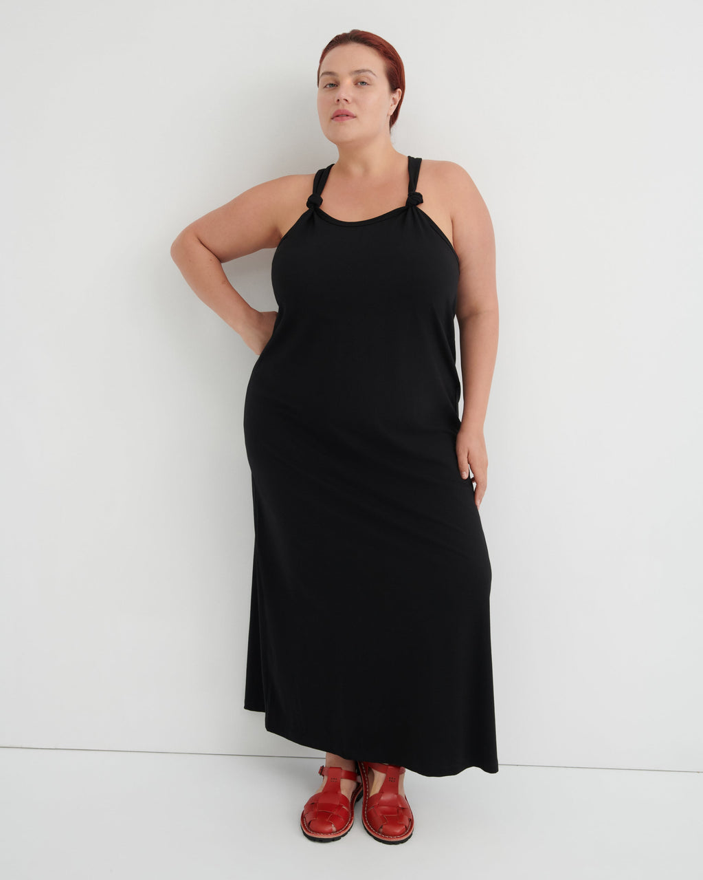 A woman modeling a black sleeveless maxi dress with thin shoulder straps and red shoes, standing against a plain white studio background.