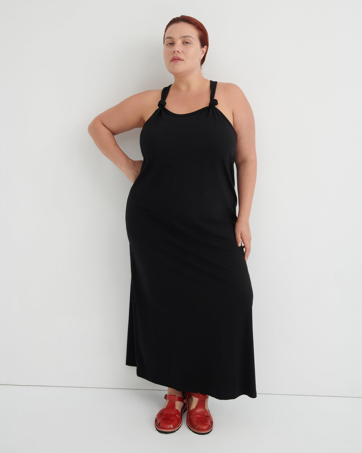 A woman modeling a black sleeveless maxi dress with thin shoulder straps and red shoes, standing against a plain white studio background.
