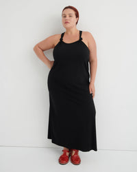 A woman modeling a black sleeveless maxi dress with thin shoulder straps and red shoes, standing against a plain white studio background.