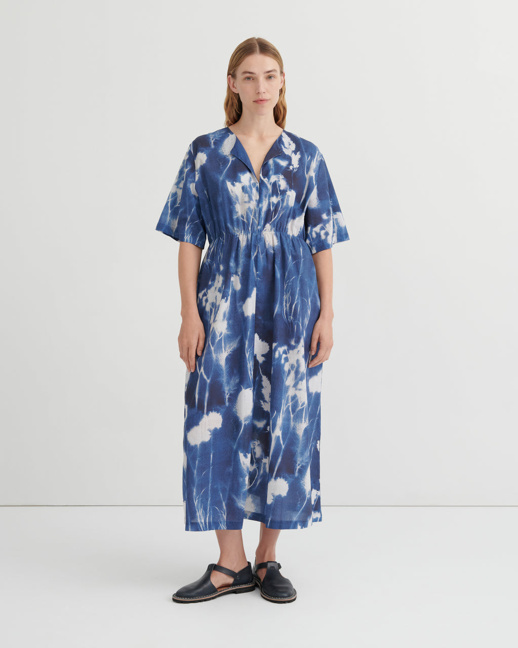 A woman stands against a plain light gray studio background wearing a loose, short-sleeve midi dress with a blue and white floral cyanotype print. The dress has a V-neck and gathered waist.