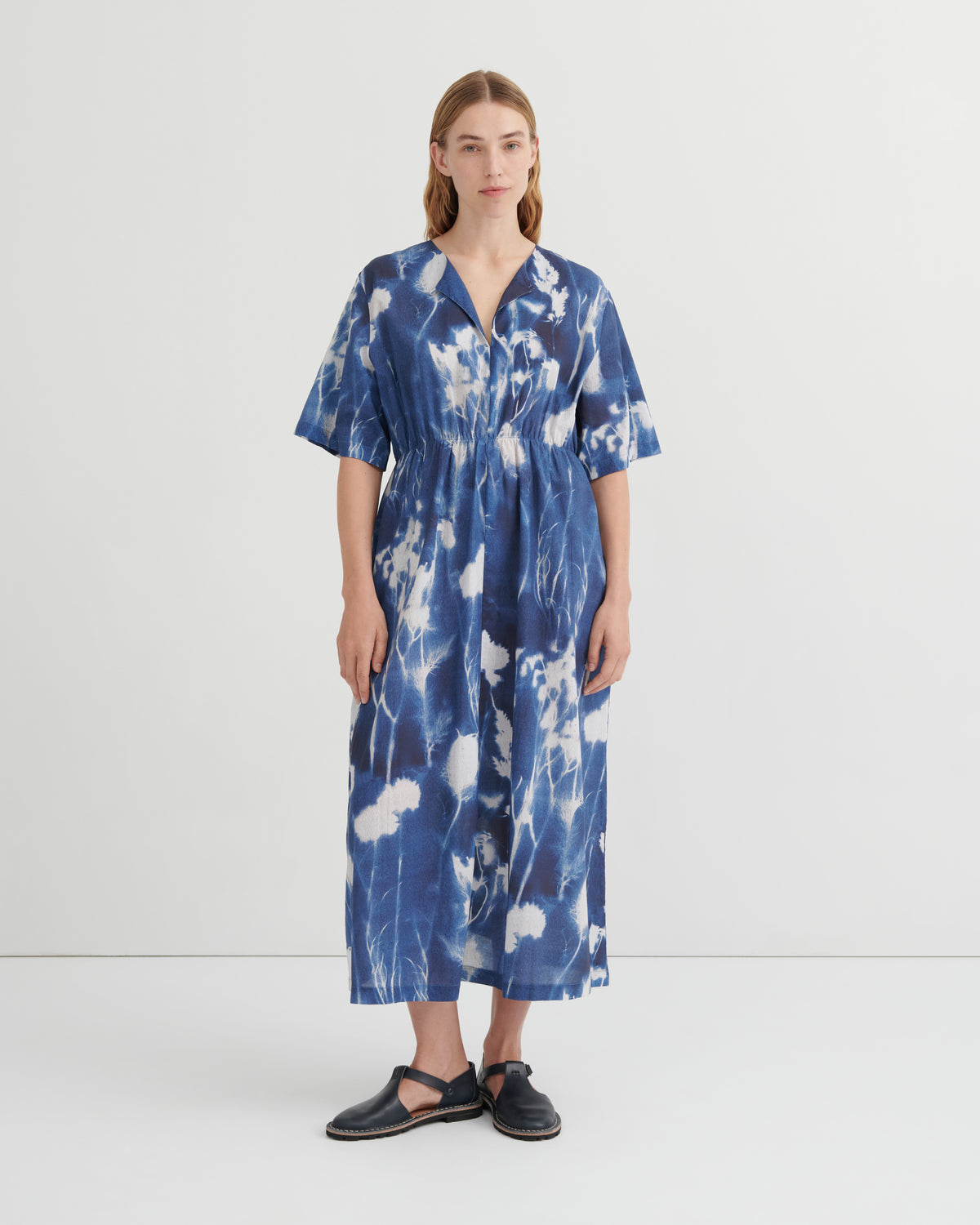 A woman stands against a plain light gray studio background wearing a loose, short-sleeve midi dress with a blue and white floral cyanotype print. The dress has a V-neck and gathered waist.