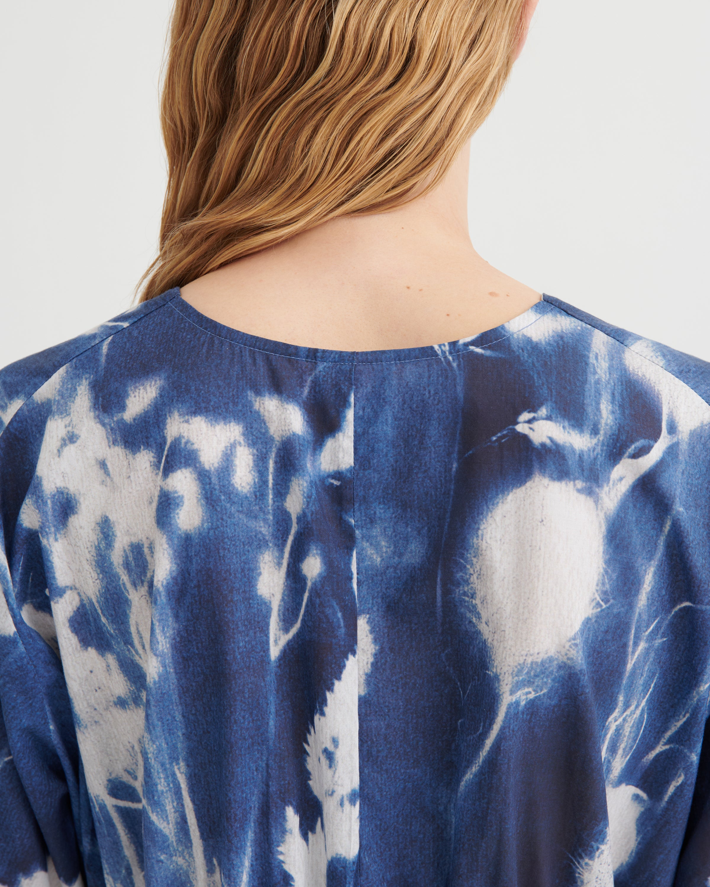 Close-up back view of a person wearing a loose blue and white tie-dye top against a plain light background.