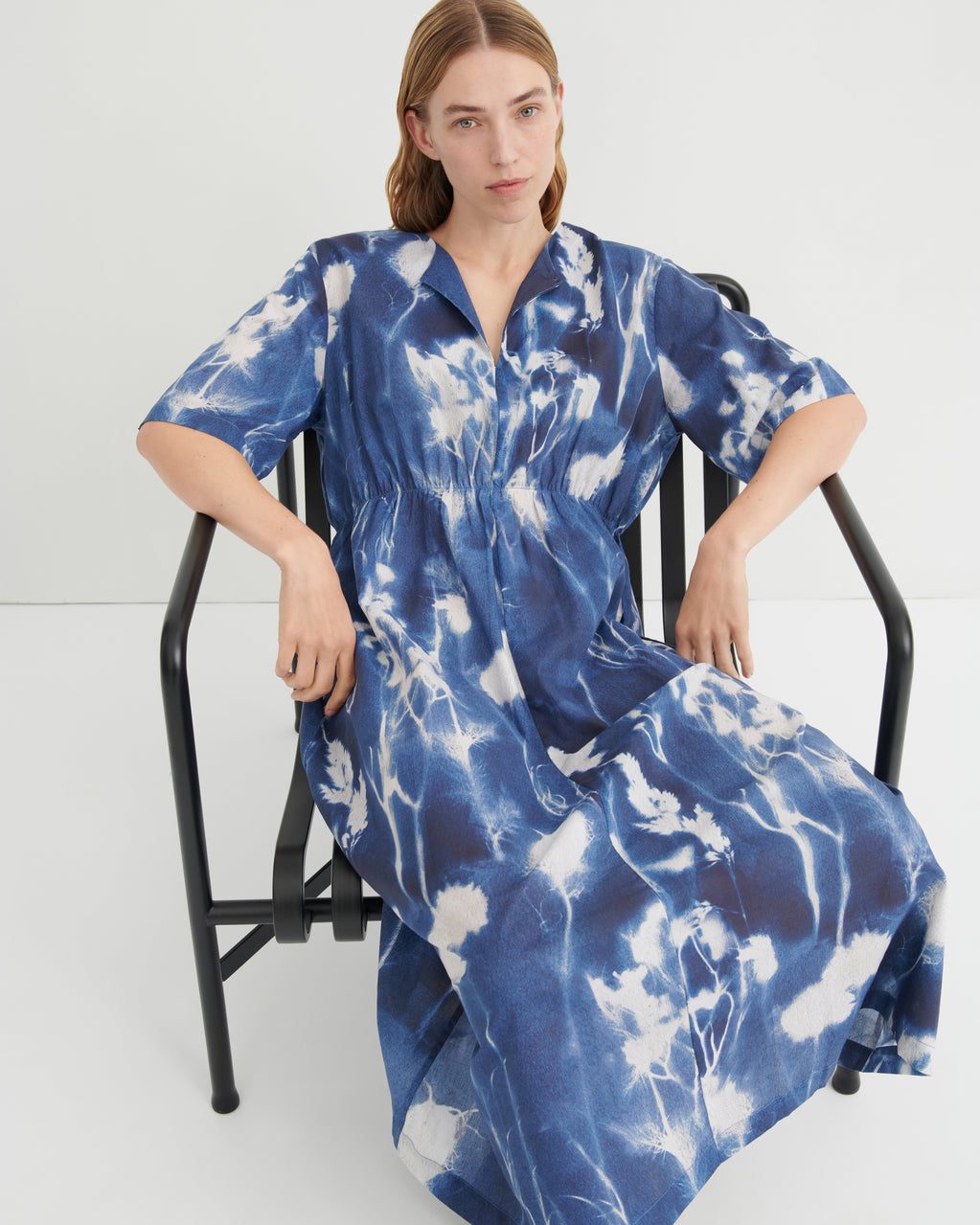 A woman wearing a blue and white tie-dye kaftan dress sits on a black metal chair against a plain white studio background.