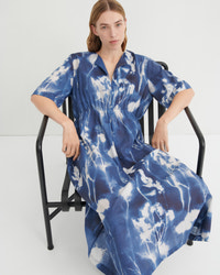 A woman wearing a blue and white tie-dye kaftan dress sits on a black metal chair against a plain white studio background.