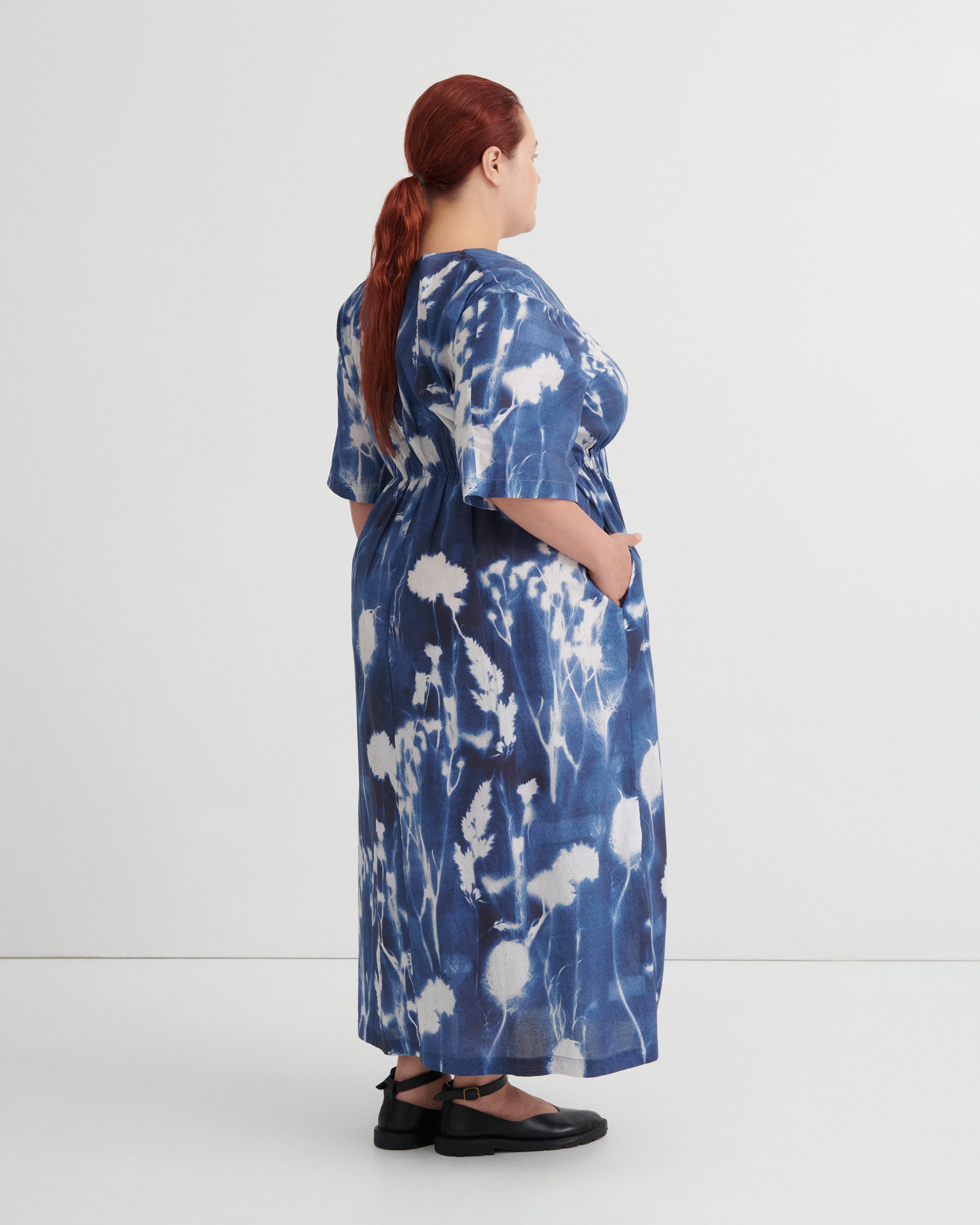 A woman with red hair tied back in a ponytail is shown from the back wearing a blue and white abstract patterned maxi dress with short wide sleeves and black flat shoes, standing against a plain light gray studio background.