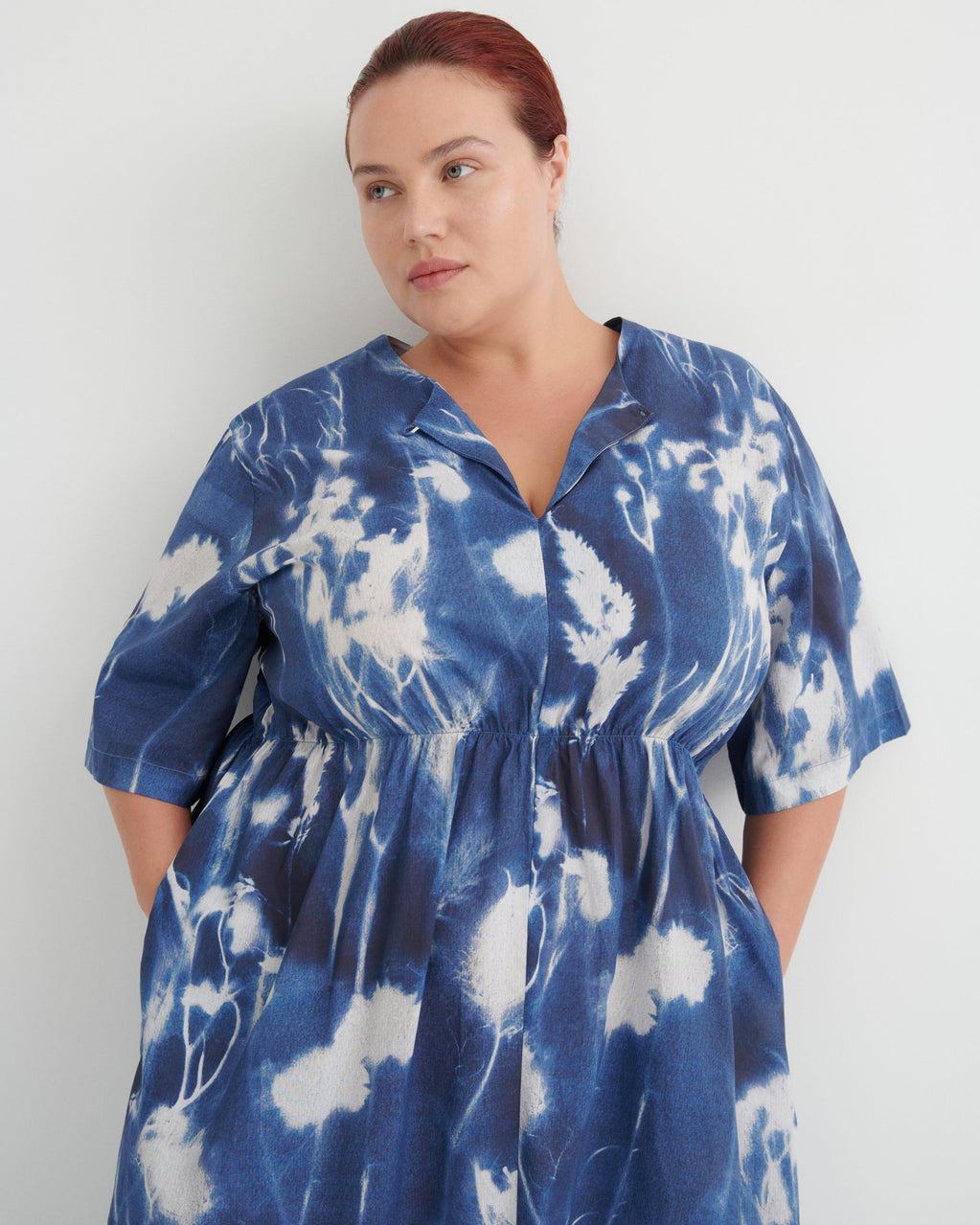 A woman wearing a blue and white tie-dye short-sleeve shirt dress, standing against a plain light gray wall.