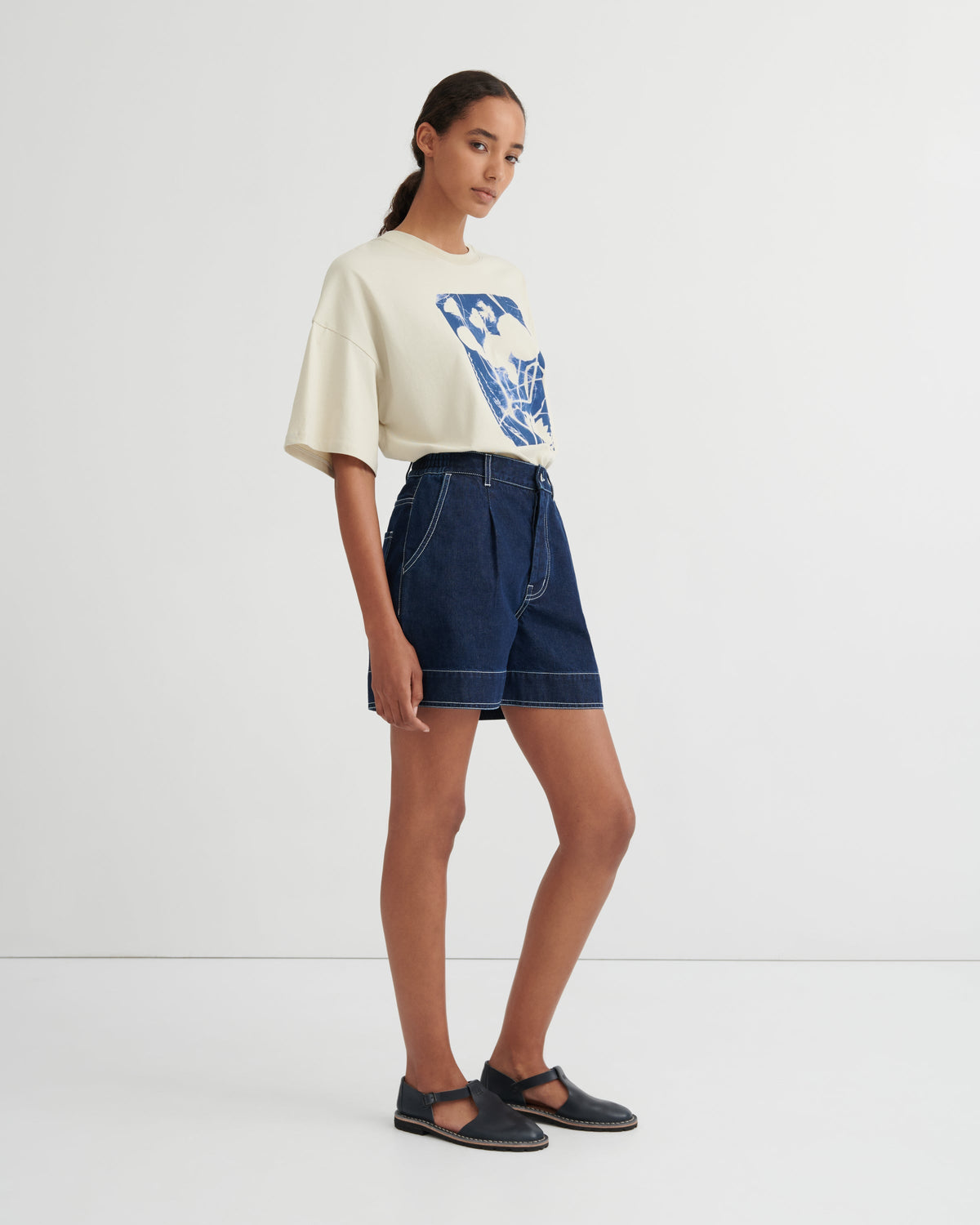 A model stands in a studio wearing a cream-colored oversized t-shirt with a blue graphic print, dark blue denim shorts, and dark navy Mary Jane flats.