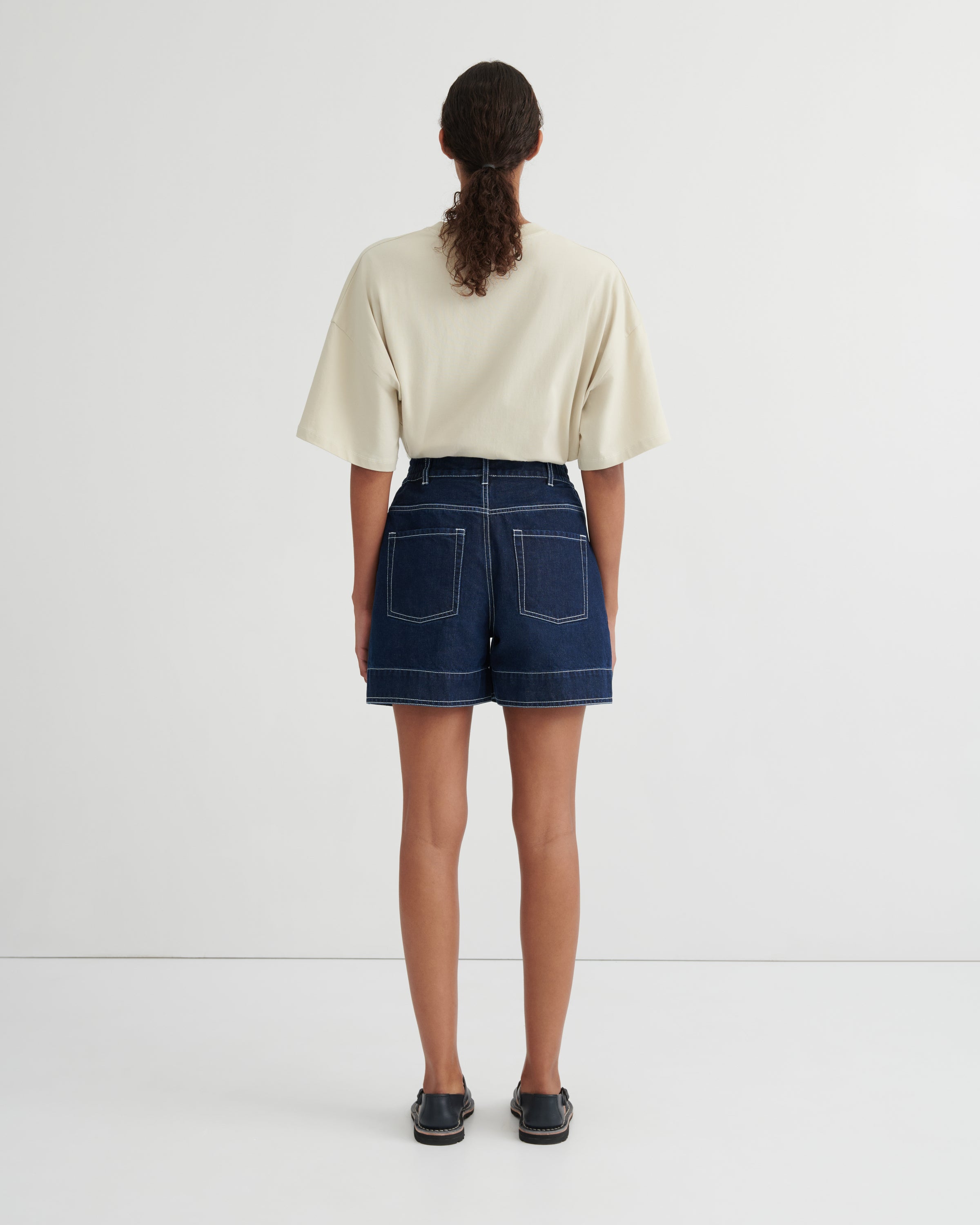 A model viewed from behind wearing a cream-colored short-sleeve crop top and dark blue denim shorts with contrast stitching, paired with dark loafers, against a plain white studio background.