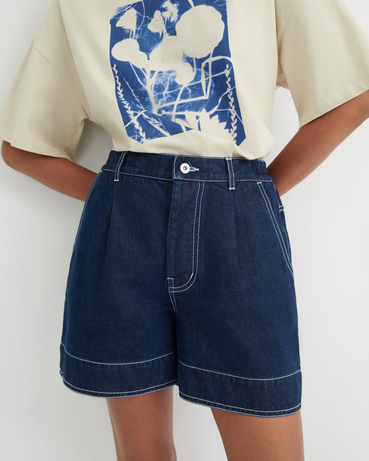 A person wearing dark blue denim shorts with contrast white stitching and a cream graphic T-shirt, shown from the chest to the upper thighs against a plain light background.