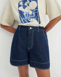 A person wearing dark blue denim shorts with contrast white stitching and a cream graphic T-shirt, shown from the chest to the upper thighs against a plain light background.