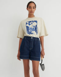 A model stands against a light gray studio background wearing a cream-colored oversized graphic T-shirt with a blue abstract print and dark blue denim shorts. She is holding a small gardening trowel in her right hand.