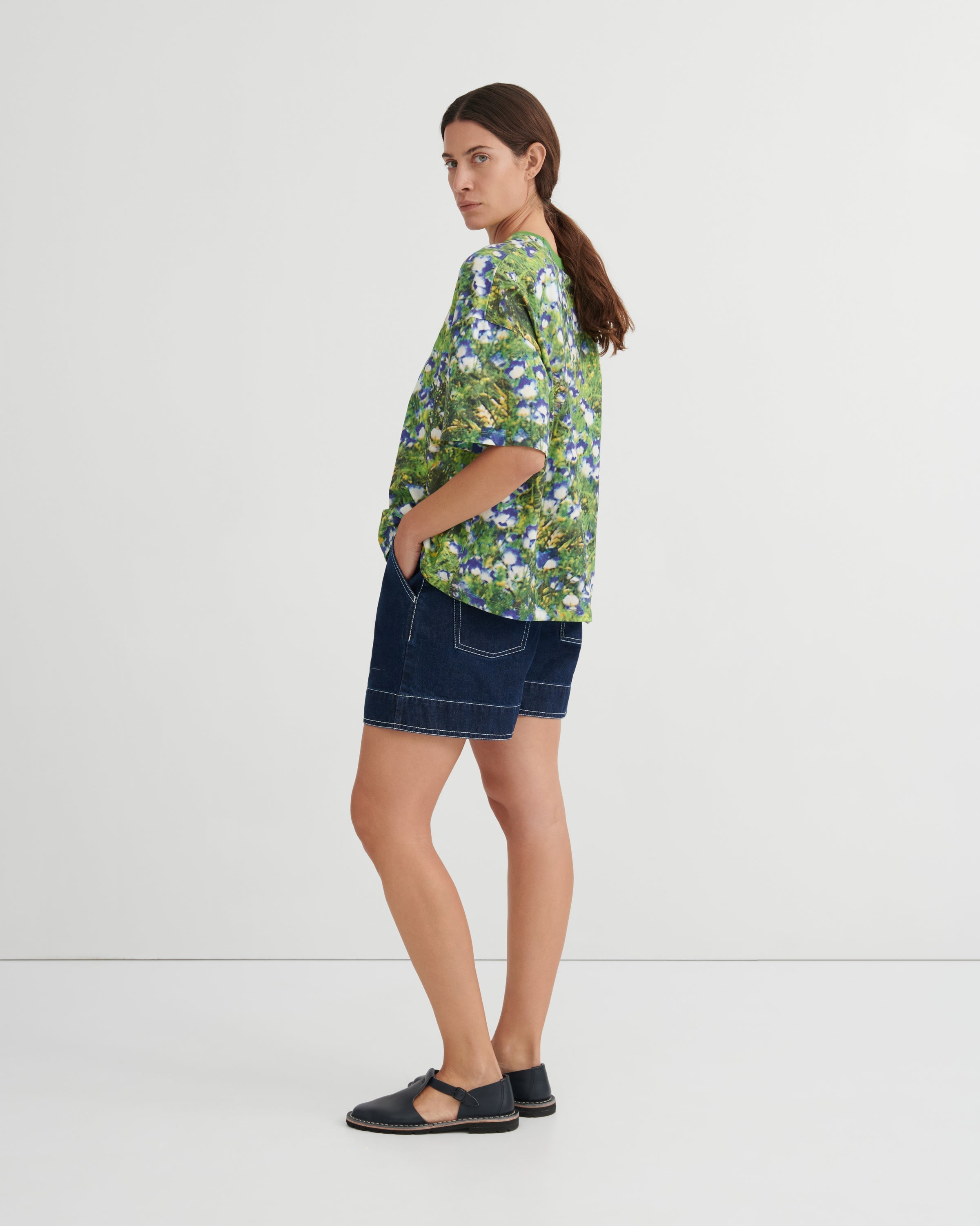 A woman stands in profile wearing a green short-sleeve button-up shirt with a blue and white floral print, dark denim shorts, and black flat sandals against a plain light gray studio background.