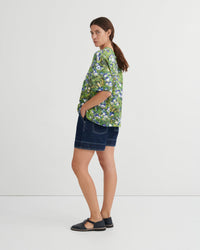A woman stands in profile wearing a green short-sleeve button-up shirt with a blue and white floral print, dark denim shorts, and black flat sandals against a plain light gray studio background.