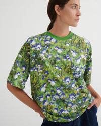 A woman wearing a short-sleeve crewneck t-shirt with a green and blue floral print, shown from the side against a plain light gray background.