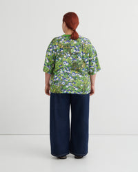 A person viewed from the back wearing a green short-sleeve top with a dense blue and white floral print, paired with dark wide-leg pants, against a plain light gray studio background.