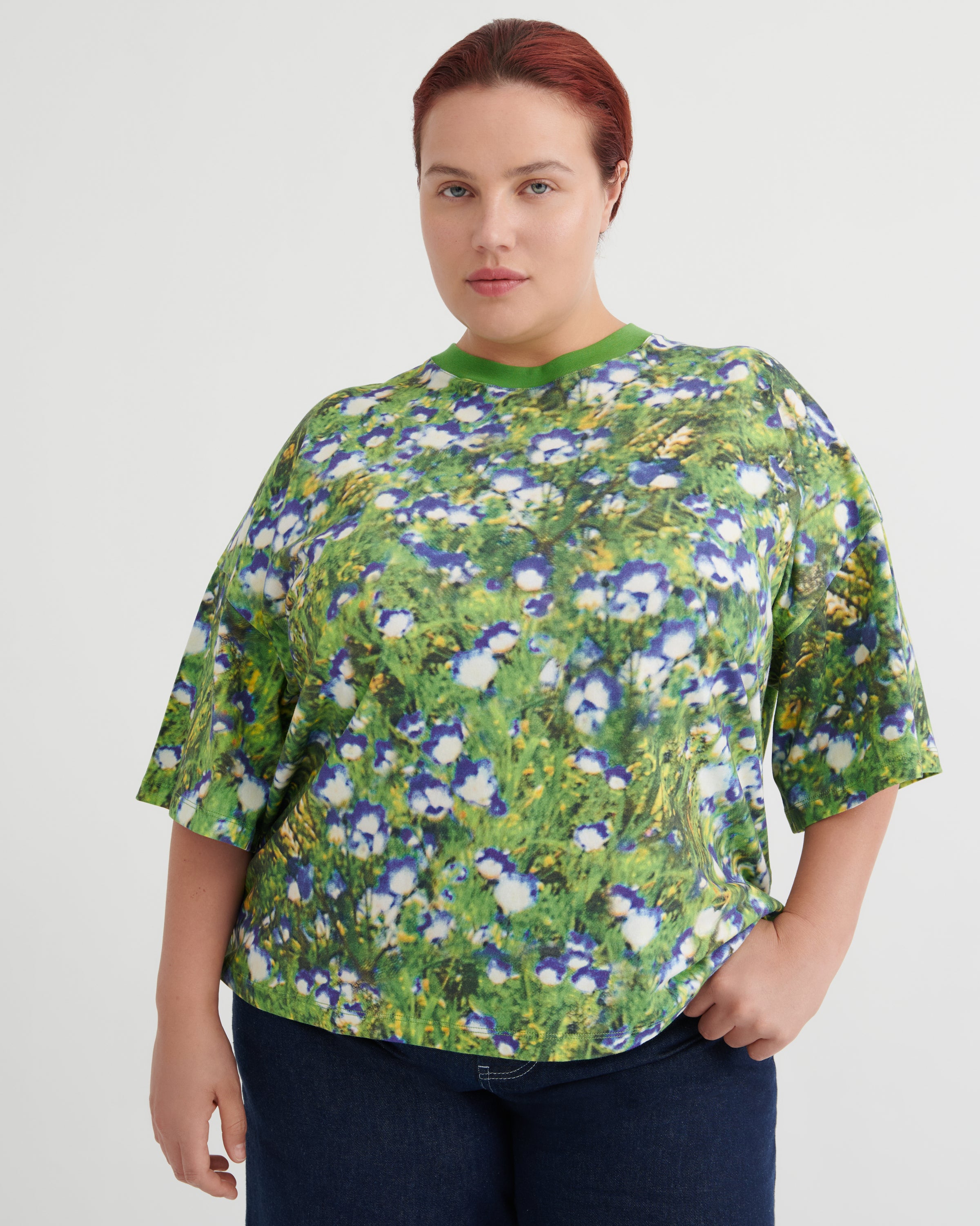 A woman wearing a short-sleeve crewneck top with a green, blue, and white floral print, paired with dark blue jeans, against a plain light gray background.