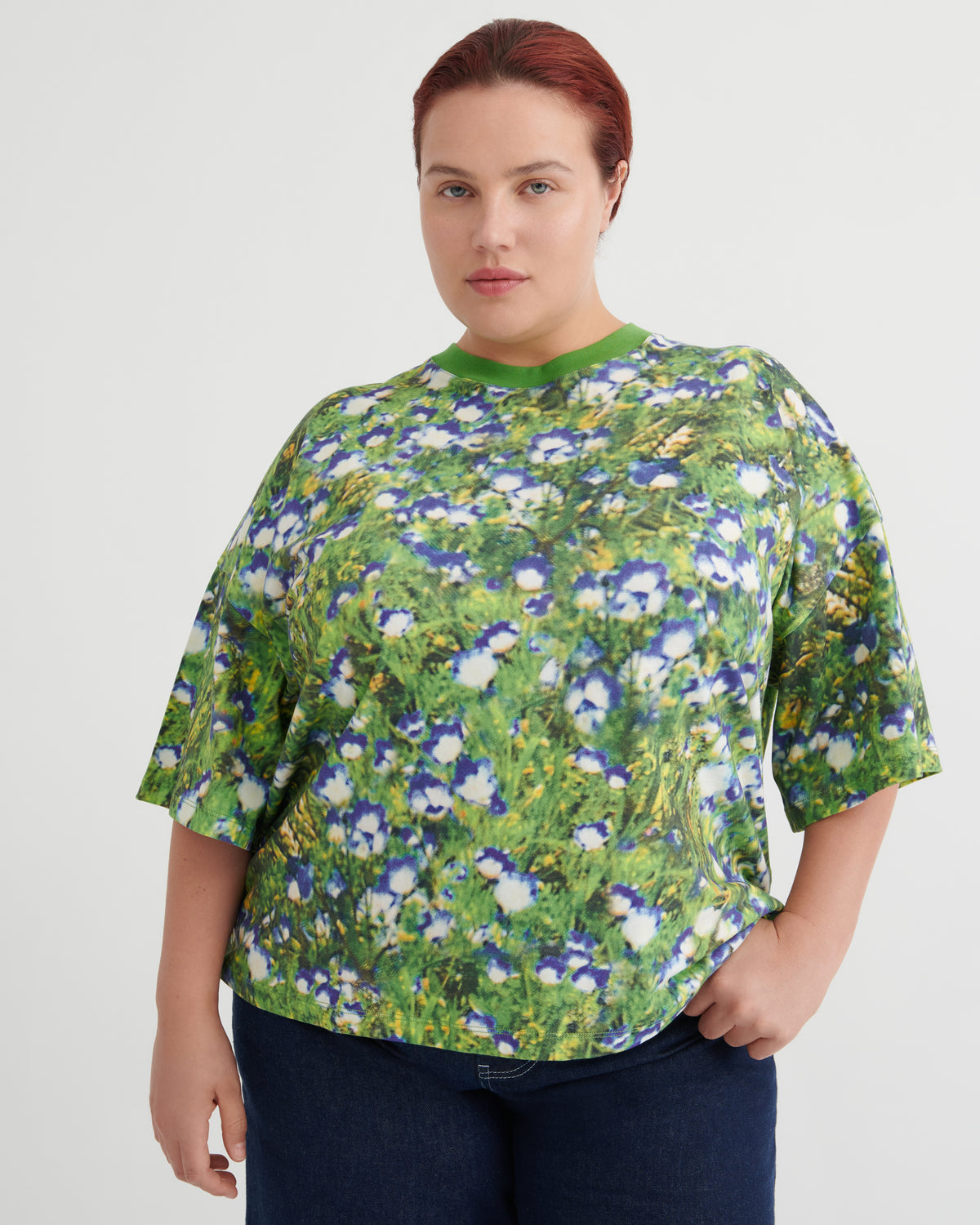 A woman wearing a short-sleeve crewneck top with a green, blue, and white floral print, paired with dark blue jeans, against a plain light gray background.